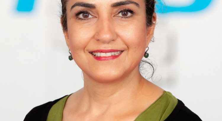Das Bild zeigt Ayşe Demir. Sie steht vor einem TBB-Logo im Hintergrund. Sie hat schwarze, auf dem Bild zusammengebundene Haare. Sie trägt eine schwarze Strickjacke über einem günen T-Shirt, Ohrringe und einen Stecker im Nasenflügel sowie Lippenstift und dezentes Augen-Make-up.